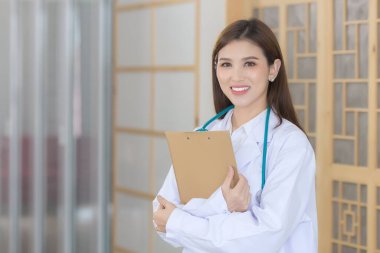 Genç ve güzel Asyalı kadın doktor kollarını kavuşturup mutlu mesut ayakta duruyor ve hastanede gülümsüyor. Beyaz bir bornoz ve steteskop giyiyordu..