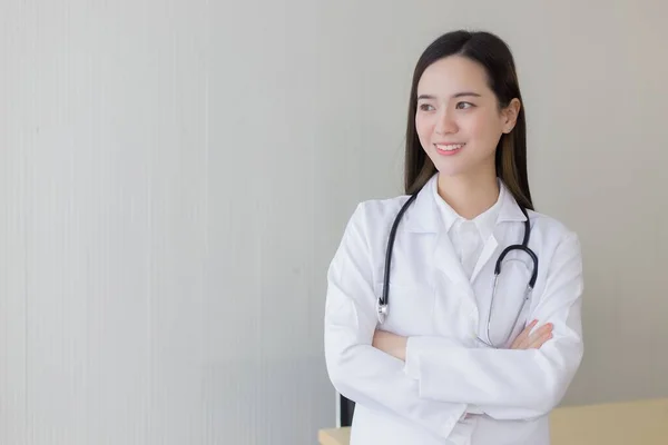 黒い長い髪のアジアの女性医師は 白いラボのコートと聴診器を身に着けています 彼女は病院で腕を組んで — ストック写真