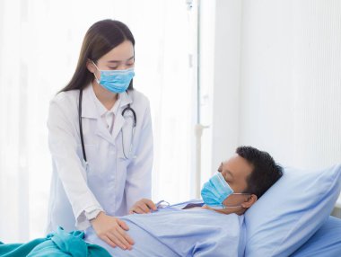 Asyalı bir kadın doktor hastanede yatan bir hastanın semptomlarını kontrol ediyor. Her ikisi de Corona virüsünü korumak için cerrahi maske takıyor (Covid 19).
