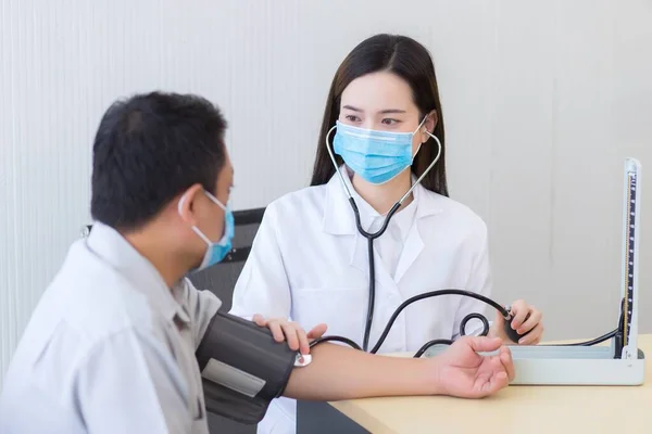 アジアの女性医師は 血圧計の男性患者とポンプゴムの折り畳み式の関節に置かれた聴診器を使用してパルス 心拍数 を聞きます 二人とも病院で顔マスクをして — ストック写真