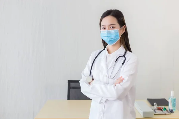 黒い長い髪をしているアジア系女性の医師は 医療用フェイスマスクを着用し 病院のオフィスで腕を組んでいる間 一様として聴診器と白衣を着用する 病院で医療面マスクを着用する — ストック写真
