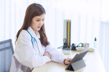 Coronavirus koruma konsepti. Asyalı ciddi profesyonel doktor beyaz önlük ve steteskop giyiyor. Modern dokunmatik ekran aletini iş yerinde dijital tablet bilgisayar kullanıyor..