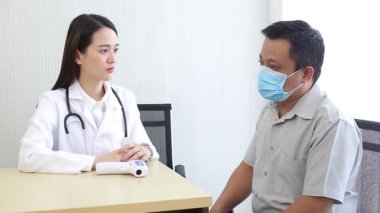 Tıbbi maske takan Asyalı bir erkek hasta vücudunda bir yerin ağrıdığını açıklarken bir kadın doktor ona hastanede bazı bilgiler veriyor..