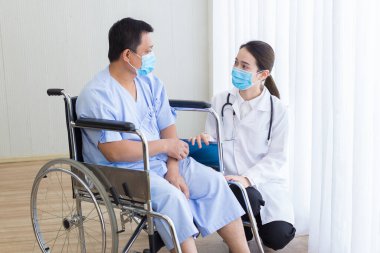 Asyalı kadın doktor, tekerlekli sandalyeye oturan bir hastaya bazı bilgiler verdi. Hastanede konuşurken tıbbi maske takıyorlar..