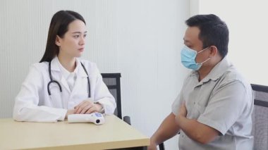 Tıbbi maske takan Asyalı bir erkek hasta vücudunda bir yerin ağrıdığını açıklarken bir kadın doktor ona hastanede bazı bilgiler veriyor.