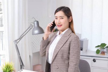 Resmi takım elbiseli kendine güvenen Asyalı iş kadını modern ofis masasında akıllı telefonla konuşuyor liderlik, iletişim ve profesyonel başarıyı simgeliyor..