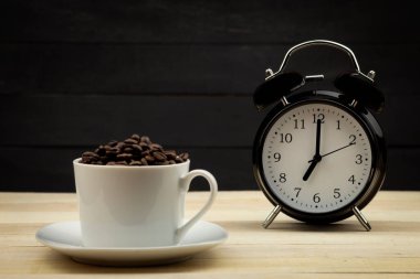 Alarm saati ve kavrulmuş kahve çekirdekleri arka plan olarak beyaz bir fincanda. Metin Şablonu Saat sabah yediyi gösteriyor