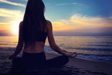 Genç bir kadının lotus poz sahil günbatımı bir arka plan üzerinde yoga meditasyon yaparken arkadan görünüm.