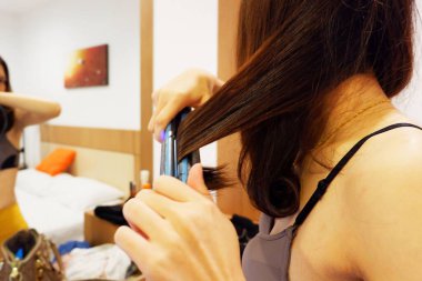 Asyalı kadın saçlarını makineden yapılma bir aletle sallıyor ve yatak odasında aynanın önünde duruyor..