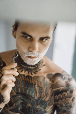 bir ayna, onun elinde bir tıraş jilet önünde genç bir yakışıklı dövmeli adam sabah tıraş.