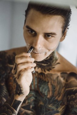 bir ayna, onun elinde bir tıraş jilet önünde genç bir yakışıklı dövmeli adam sabah tıraş.