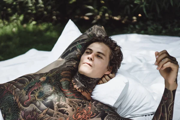 a young handsome tattooed man in the morning in bed tosses up the pillows. bed in the garden