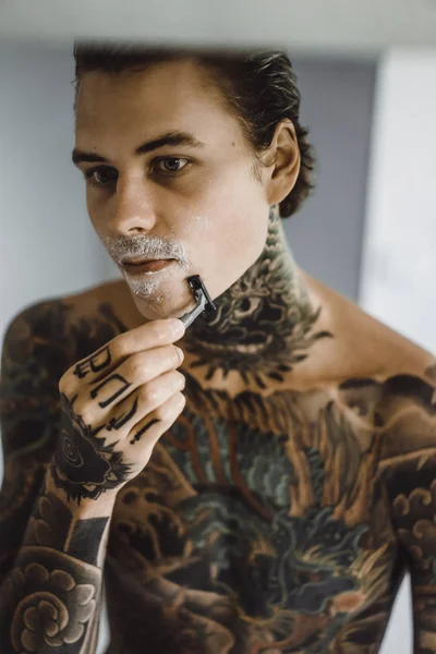 手剃りのカミソリ 鏡の前で朝の若いハンサムな刺青男がひげそり — ストック写真