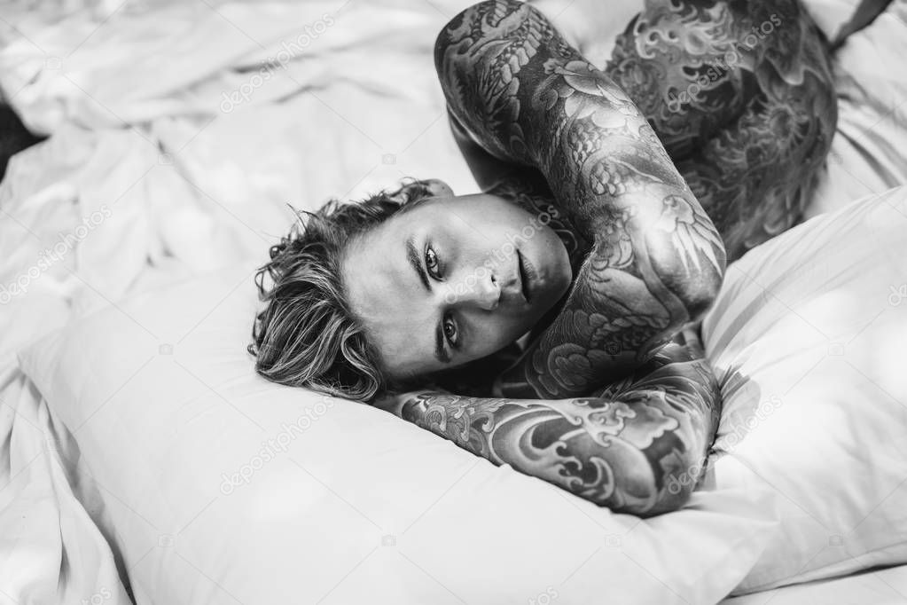 Handsome young tattooed man relaxing on white bed