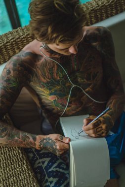 dövmeler kulaklıklar giyen genç bir erkekte müziği dinliyor ve içinde a defter bir kalem çizer. Çevrimiçi eğitim.