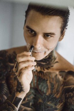 bir ayna, onun elinde bir tıraş jilet önünde genç bir yakışıklı dövmeli adam sabah tıraş.