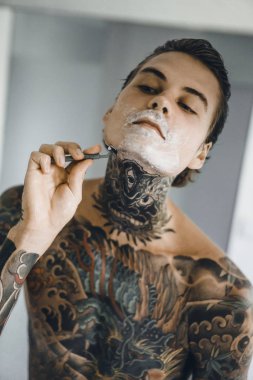 bir ayna, onun elinde bir tıraş jilet önünde genç bir yakışıklı dövmeli adam sabah tıraş.
