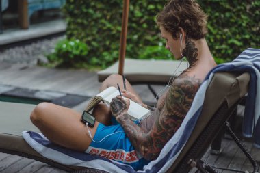 dövmeler kulaklıklar giyen genç bir erkekte müziği dinliyor ve içinde a defter bir kalem çizer. Çevrimiçi eğitim.