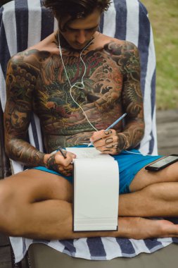 dövmeler kulaklıklar giyen genç bir erkekte müziği dinliyor ve içinde a defter bir kalem çizer. Çevrimiçi eğitim.