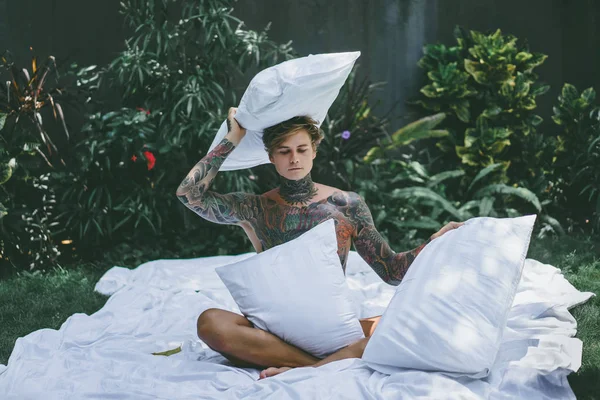 a young handsome tattooed man in the morning in bed tosses up the pillows. bed in the garden