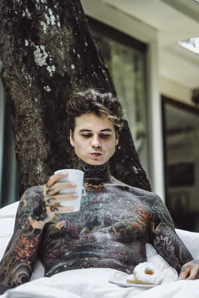 young handsome tattooed man having breakfast in bed in the open air outdoors in the garden