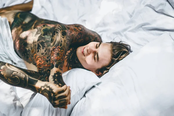 Yakışıklı genç dövmeli adam beyaz yatakta rahatlatıcı