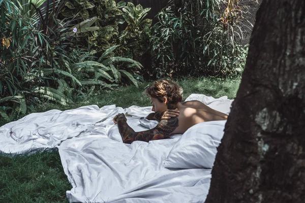 一个年轻英俊的纹身男子在早上在床上扔枕头 床在庭院里 — 图库照片