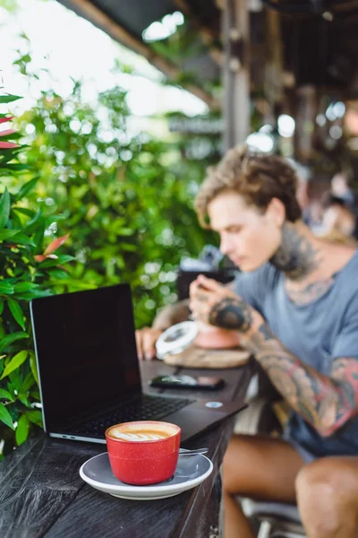 A man in tattoos breakfasts in an outdoor cafe, works on a laptop, drinks coffee. The concept of freelancing