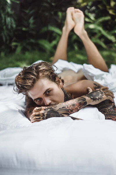 Handsome young tattooed man relaxing on white bed