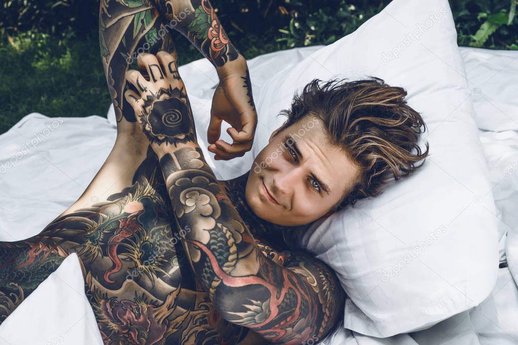 a young handsome tattooed man in the morning in bed tosses up the pillows. bed in the garden