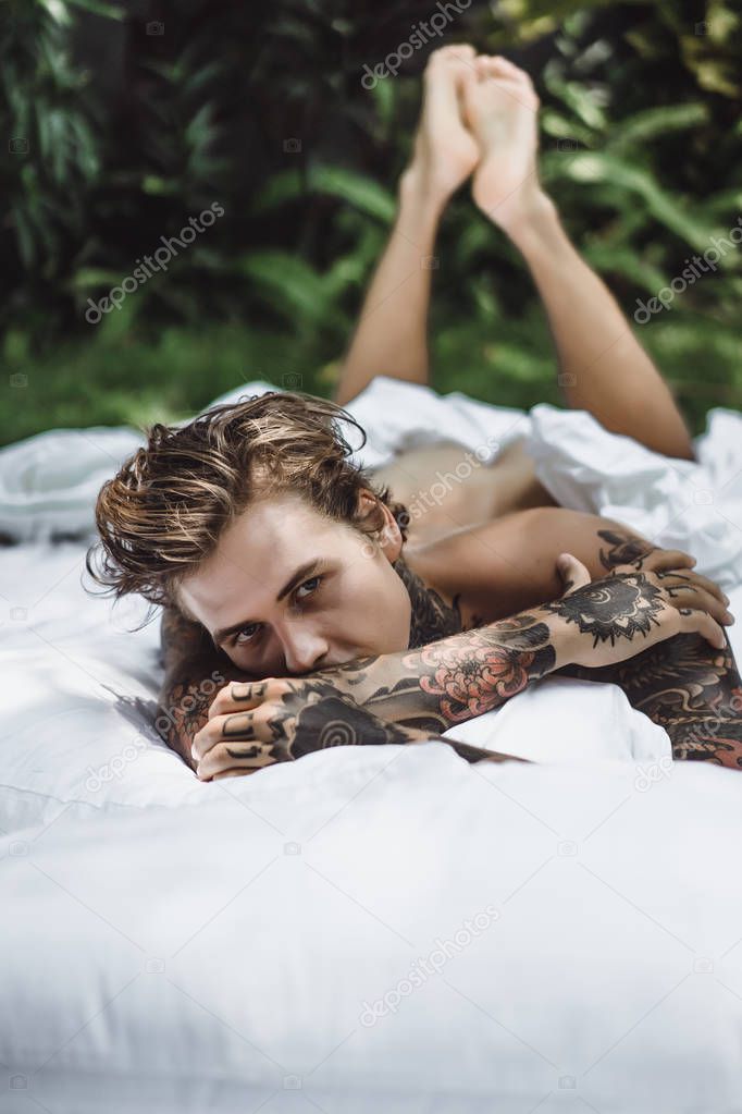 Handsome young tattooed man relaxing on white bed