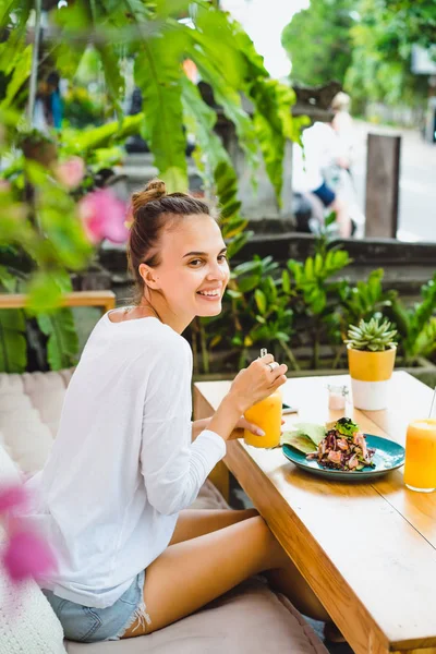 A beautiful woman has breakfast in a stylish cafe, a healthy breakfast, fruit, freshly squeezed juice, tropical location.