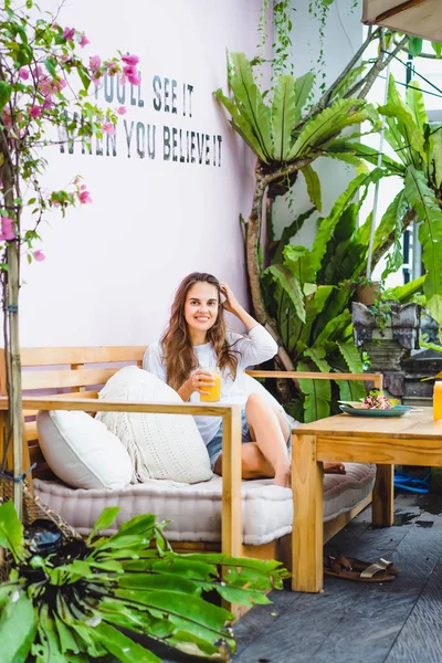 A beautiful woman has breakfast in a stylish cafe, a healthy breakfast, fruit, freshly squeezed juice, tropical location.