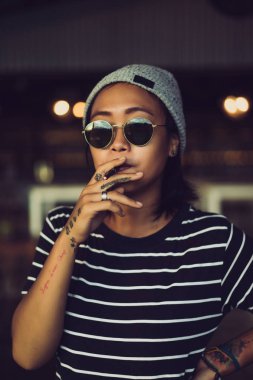 Endonezya kız hippi gri bir şapka ve güneş gözlüğü. Dövmeler, güzel şık Asyalı kadın siyah sigara içiyor.