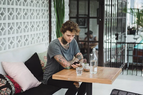 a tattooed man in a cafe uses a smartphone, drinks coffee, waiting for an order.