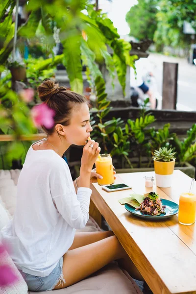 A beautiful woman has breakfast in a stylish cafe, a healthy breakfast, fruit, freshly squeezed juice, tropical location.