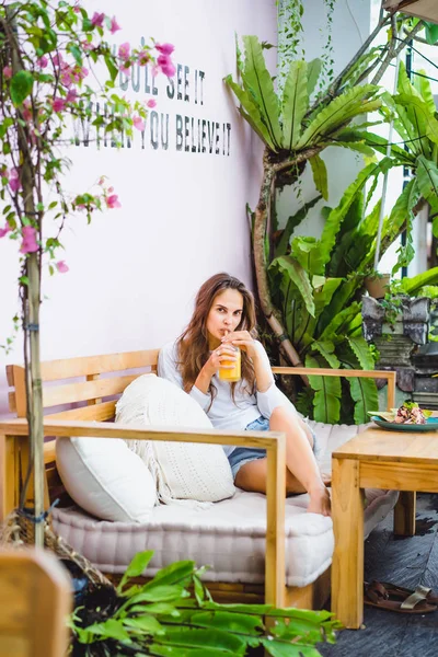 A beautiful woman has breakfast in a stylish cafe, a healthy breakfast, fruit, freshly squeezed juice, tropical location.