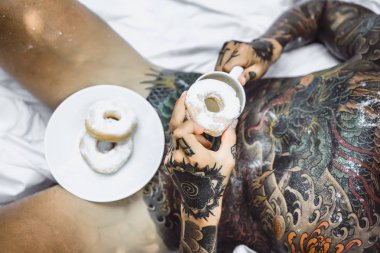 Genç, yakışıklı, dövmeli bir adam kahvaltısını yapıyor açık havada bahçede, açık havada, kahvaltıda çörek ve kahve içiyor..