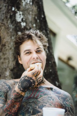 Genç, yakışıklı, dövmeli bir adam kahvaltısını yapıyor açık havada bahçede, açık havada, kahvaltıda çörek ve kahve içiyor..