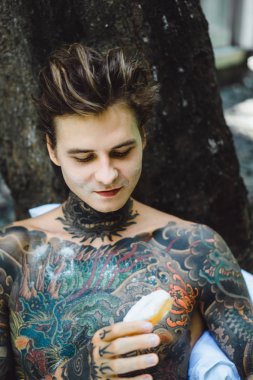 Genç, yakışıklı, dövmeli bir adam kahvaltısını yapıyor açık havada bahçede, açık havada, kahvaltıda çörek ve kahve içiyor..