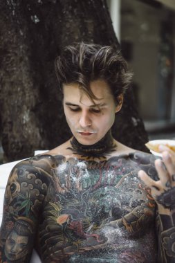 Genç, yakışıklı, dövmeli bir adam kahvaltısını yapıyor açık havada bahçede, açık havada, kahvaltıda çörek ve kahve içiyor..