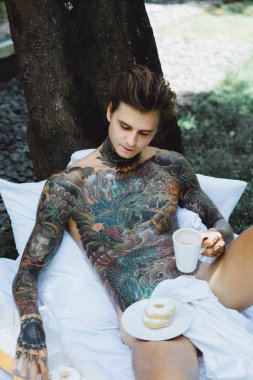 Genç, yakışıklı, dövmeli bir adam kahvaltısını yapıyor açık havada bahçede, açık havada, kahvaltıda çörek ve kahve içiyor..