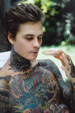 Genç, yakışıklı, dövmeli bir adam kahvaltısını yapıyor açık havada bahçede, açık havada, kahvaltıda çörek ve kahve içiyor..