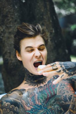 Genç, yakışıklı, dövmeli bir adam kahvaltısını yapıyor açık havada bahçede, açık havada, kahvaltıda çörek ve kahve içiyor..