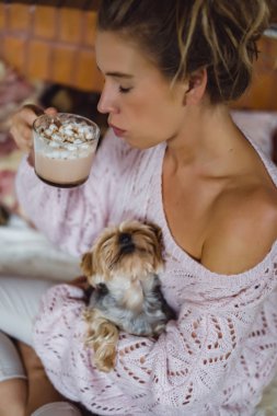 Şöminenin yanında köpeği olan genç bir kadın marşmelovlu kakao içer. Uzun saçlı güzel bir kız elinde bir York teriyeri tutuyor.