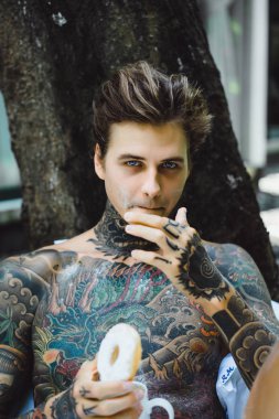 Genç, yakışıklı, dövmeli bir adam kahvaltısını yapıyor açık havada bahçede, açık havada, kahvaltıda çörek ve kahve içiyor..