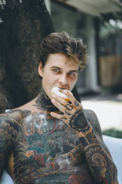 Genç, yakışıklı, dövmeli bir adam kahvaltısını yapıyor açık havada bahçede, açık havada, kahvaltıda çörek ve kahve içiyor..