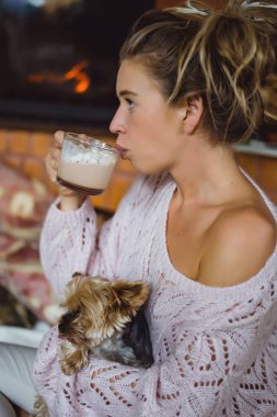 Şöminenin yanında köpeği olan genç bir kadın marşmelovlu kakao içer. Uzun saçlı güzel bir kız elinde bir York teriyeri tutuyor.