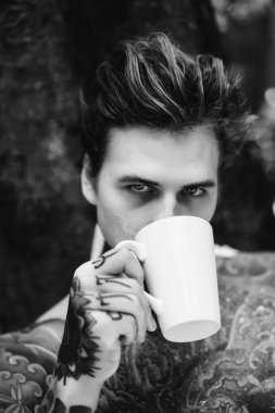 Genç, yakışıklı, dövmeli bir adam kahvaltısını yapıyor açık havada bahçede, açık havada, kahvaltıda çörek ve kahve içiyor..