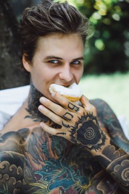 Genç, yakışıklı, dövmeli bir adam kahvaltısını yapıyor açık havada bahçede, açık havada, kahvaltıda çörek ve kahve içiyor..